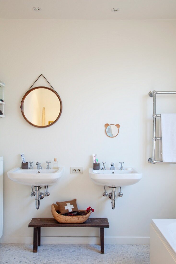 Bowl on simple wooden bench between twin sinks, round mirror on wall and partially visible stainless steel towel rack