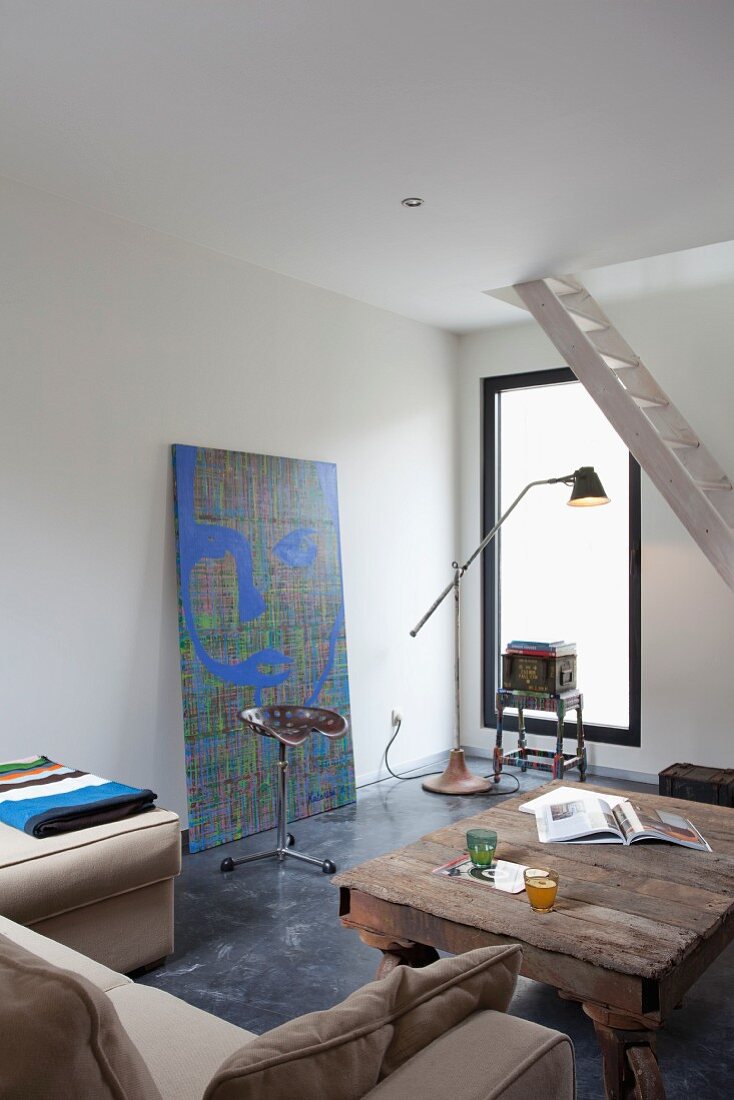 Interior with pale sofa, rustic coffee table, bar stool with saddle-style seat in front of large modern painting and ship ladder stairs in background