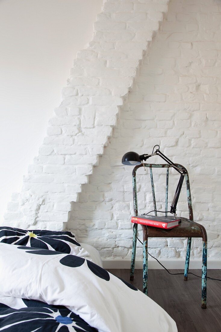 Modern, floral bed linen on partially visible bed next to retro metal chair against whitewashed brick wall
