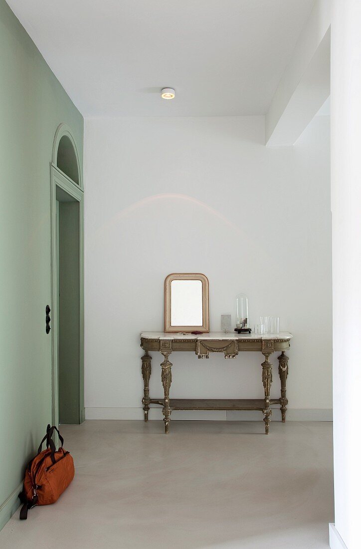 Mirror leaning against wall on antique console table in restored hallway with polished concrete floor and pastel green wall