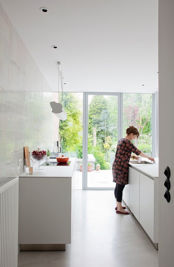 weiße Designerküche mit gefliester Wand vor geöffneter Terrassenschiebetür und Gartenblick