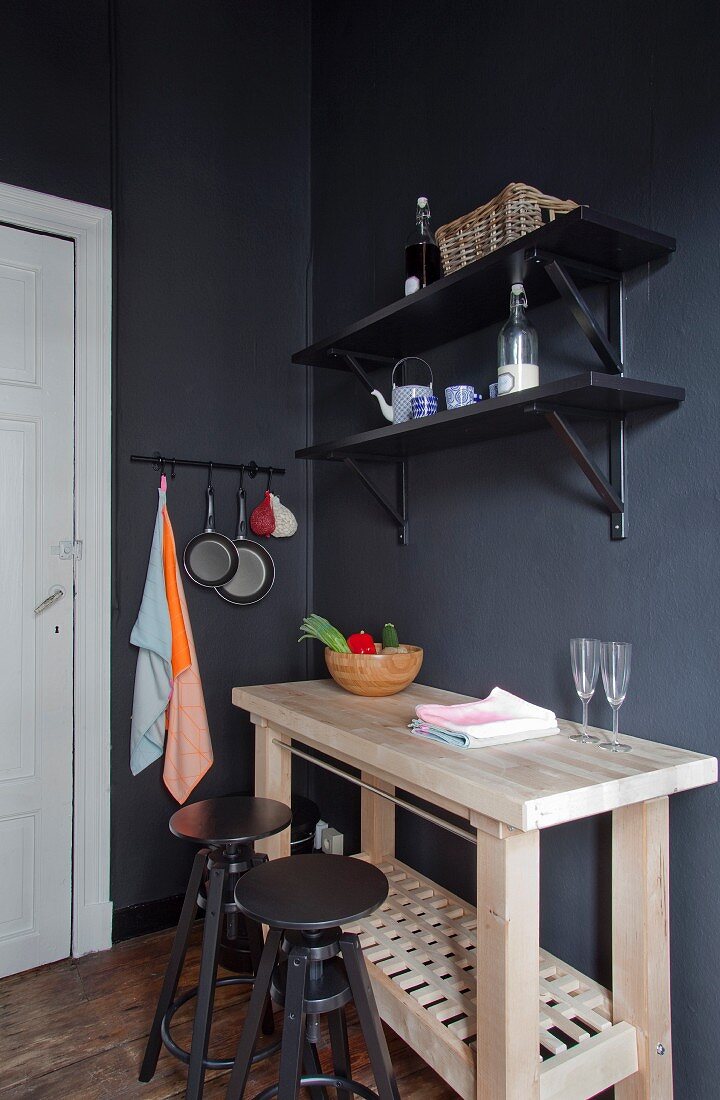 Untreated wooden kitchen counter and two dark grey bar stools below black shelves on dark wall