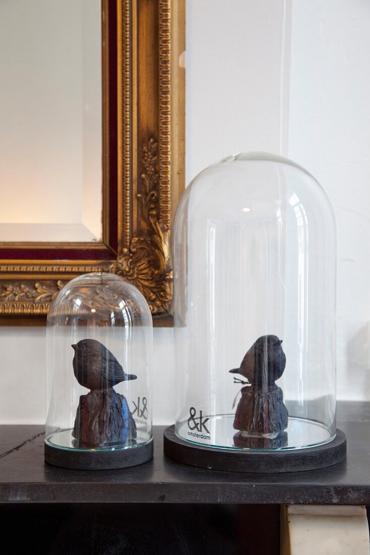 Zwei kunsthandwerkliche Vogelfiguren unter Glashauben vor Goldrahmenspiegel arrangeiert