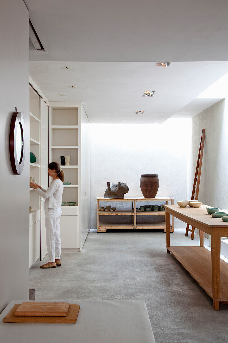 Woman in white clothing looks at objects on a shelf in a workroom