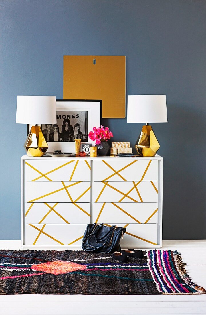 Table lamps with white lampshades and brass bases on white chest of drawers with pattern of gold stripes on drawers against wall painted blue-grey
