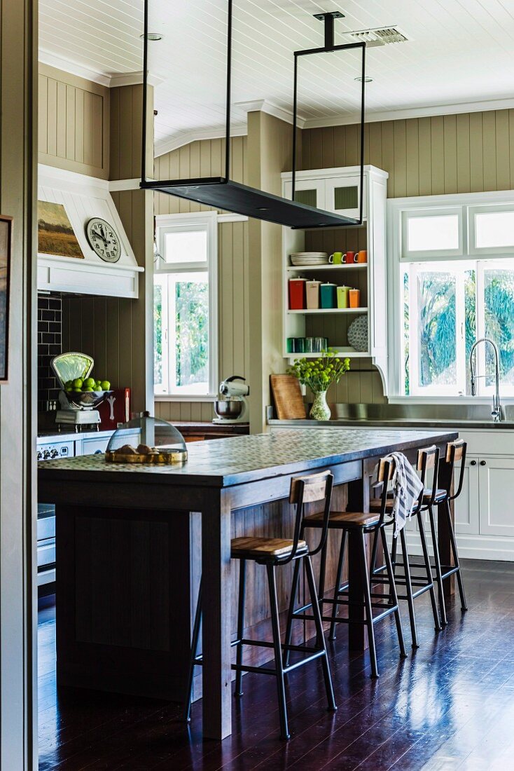 Suspended shelf over breakfast bar with bar stools on dark board floor in wood-clad kitchen with country-house charm painted light brown