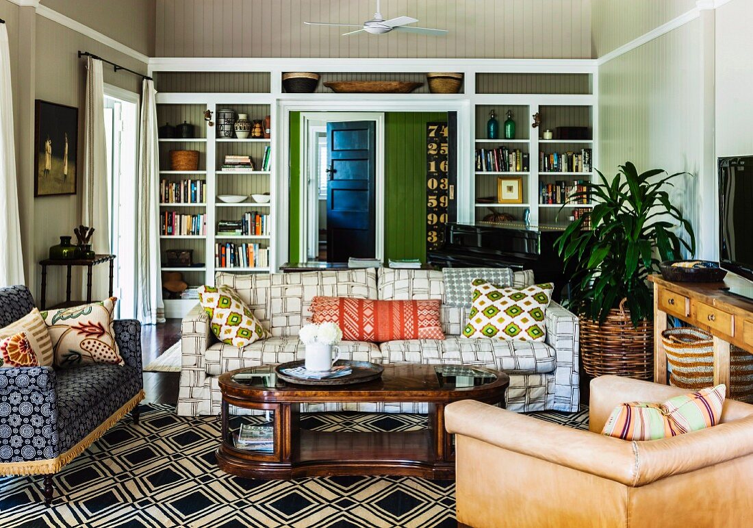 Landhaus-Wohnzimmer mit verschiedenen Polsterstühlen und gemütlichem Sofa im Mustermix um gebogenem Couchtisch aus Holz, Teppich mit schwarzweissem geometrischem Muster