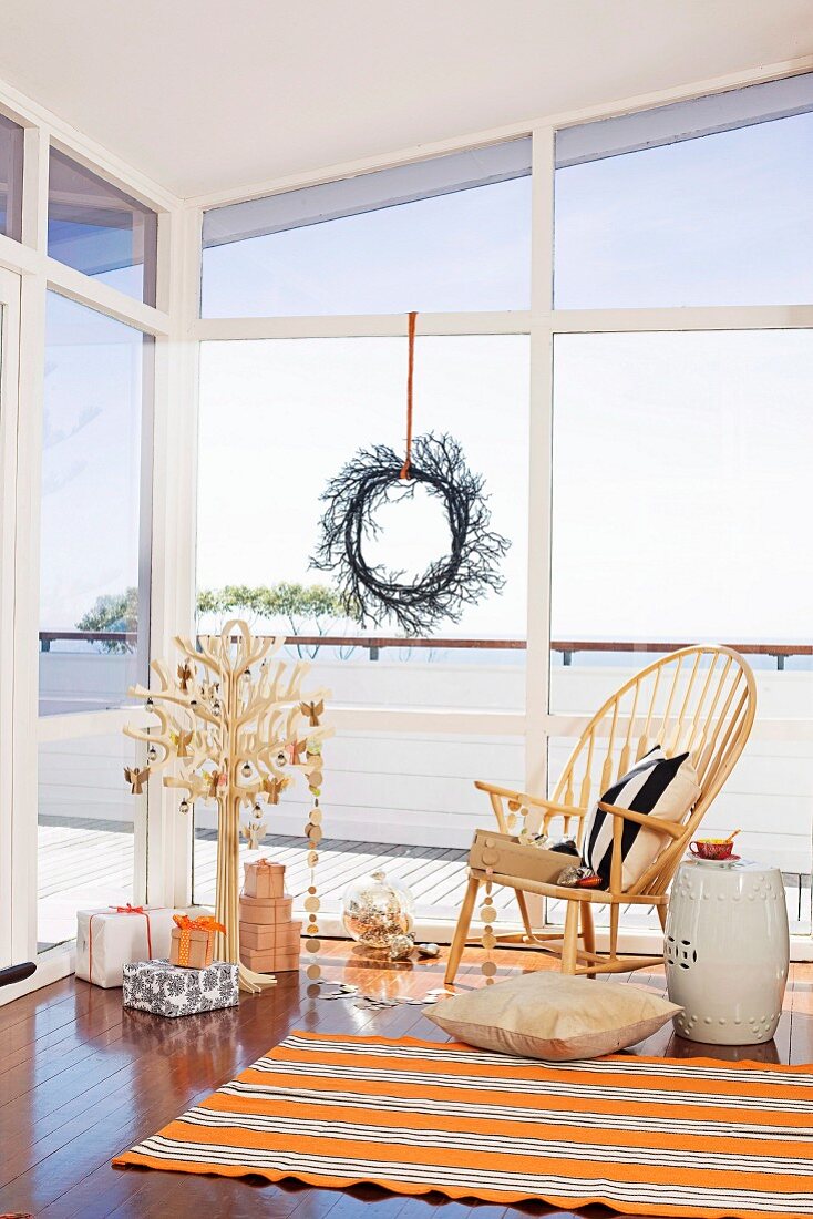 Geschenke unter stilisiertem Weihnachtsbaum und Klassikerstuhl vor Fensterfront mit Dekokranz