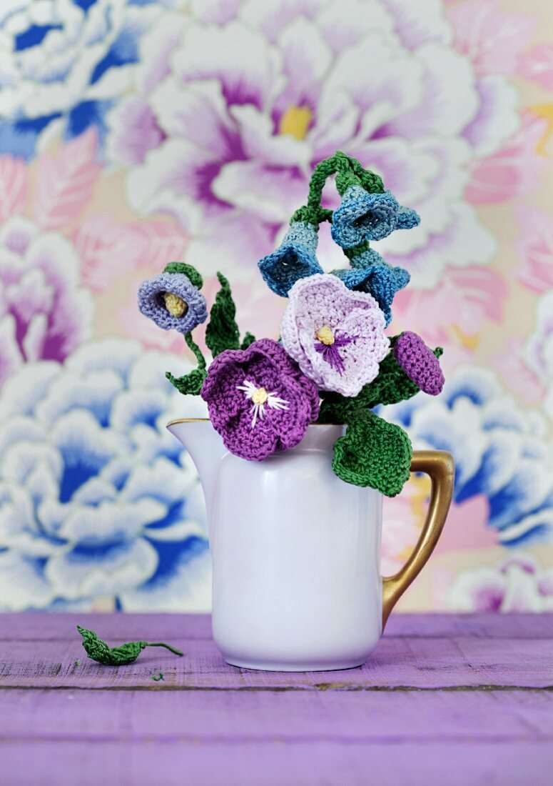 Purple, crocheted flowers in nostalgic milk jug arranged in front of floral wallpaper