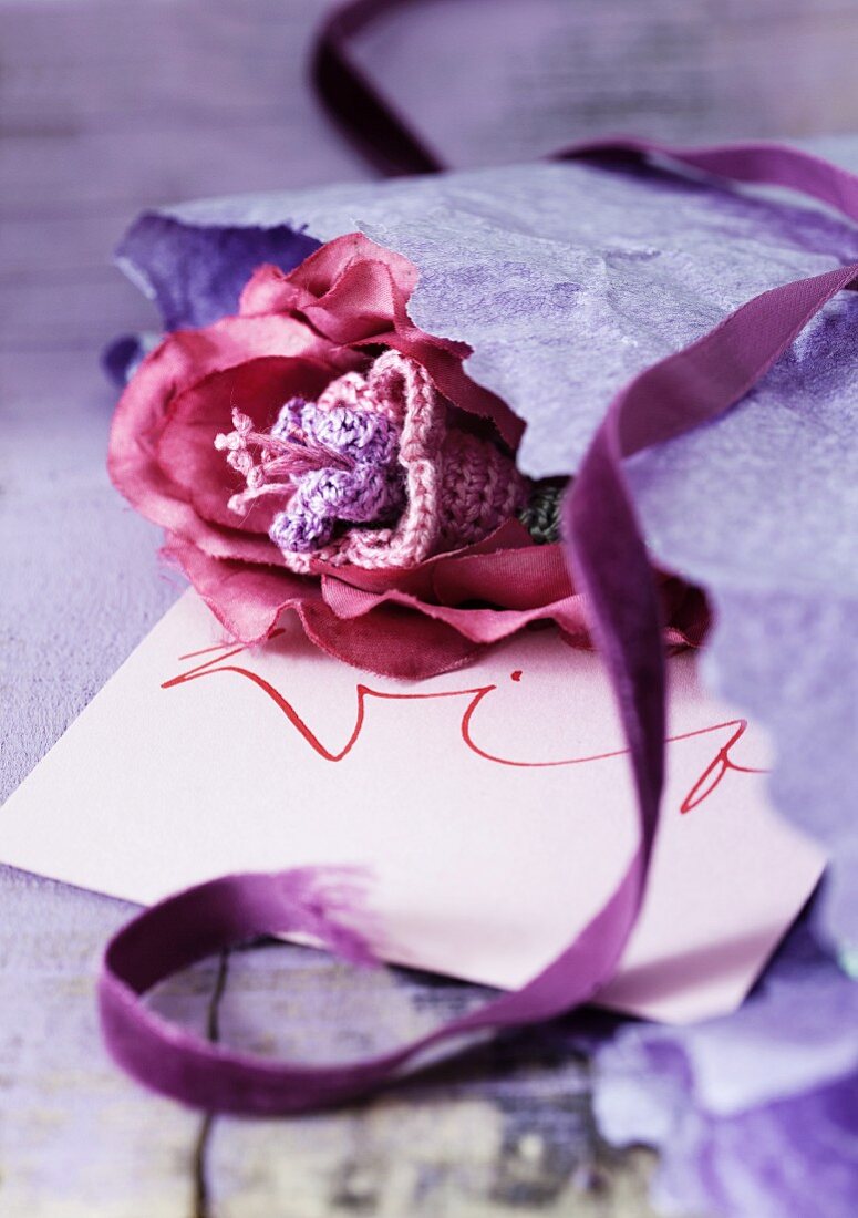 Romantische rosafarbene Häkelblüte mit Geschenkpapier, lilafarbenem Samtband und beschriftetem Briefumschlag