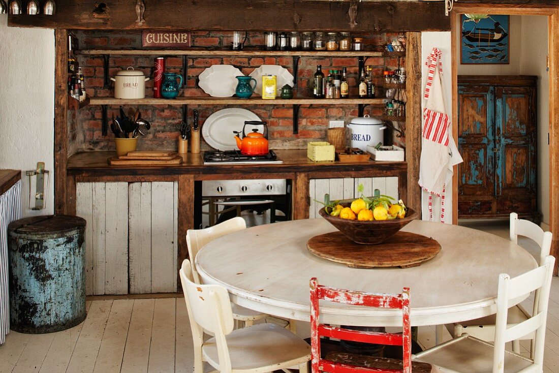 Fruit bowl on round table in front of rustic, vintage kitchen counter with open-fronted shelves