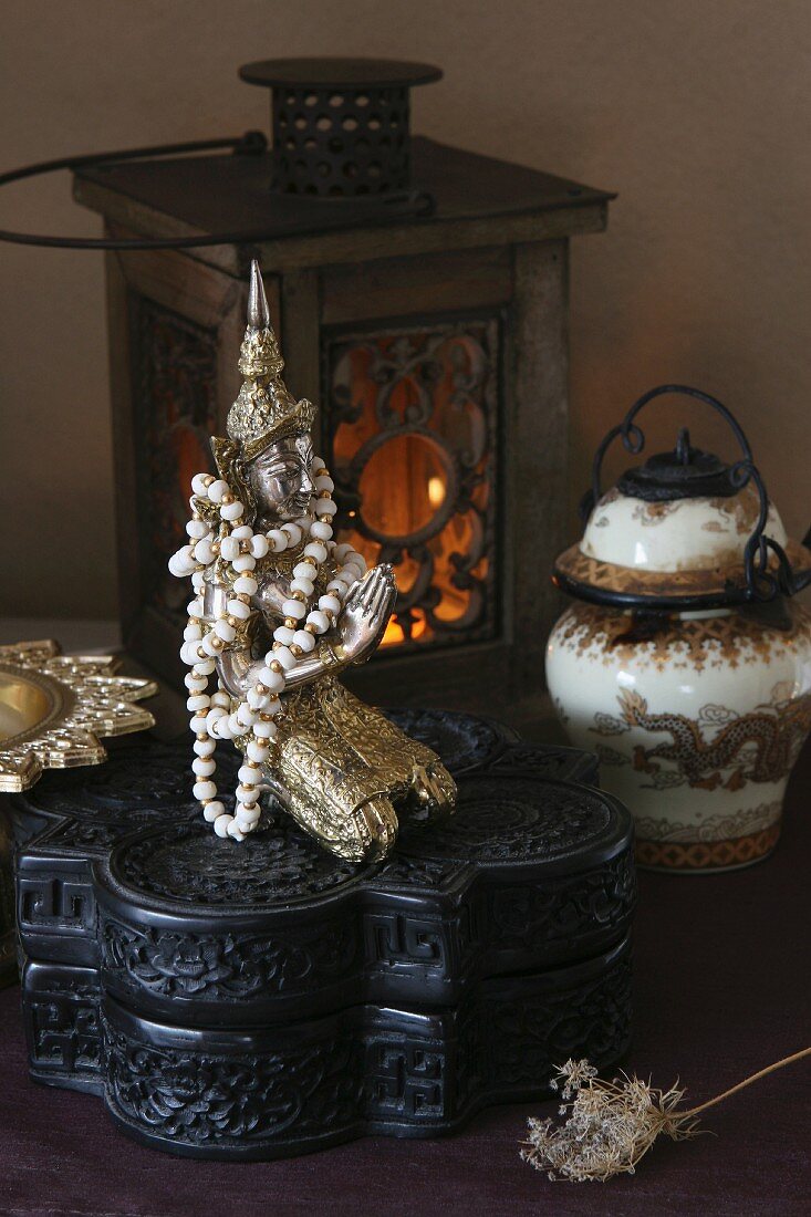 Perlenkettte um Buddha-Figur auf geschnitztem, schwarzem Holzkästchen und chinesischer Behälter vor brennender Laterne