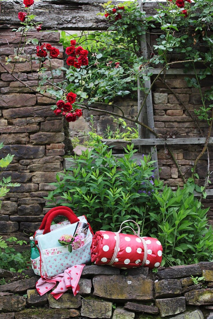 Gerollte Picknickdecke mit rotweissem Punktemuster und geblümte Picknicktasche auf Natursteinmauer vor Busch an verfallenem Haus