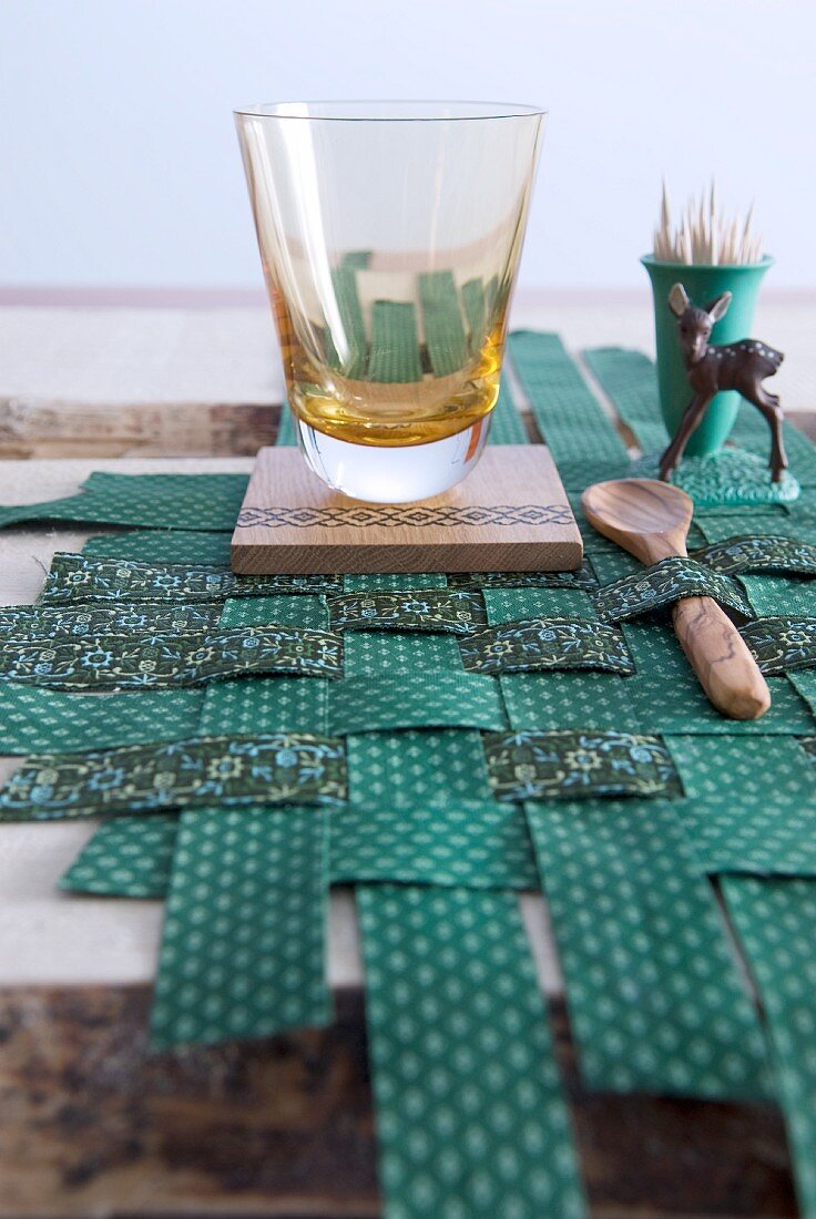 Trinkglas, Holzaccessoires und Rehfigur auf selbstgewebtem Tischset aus grün gemusterten Stoffbahnen