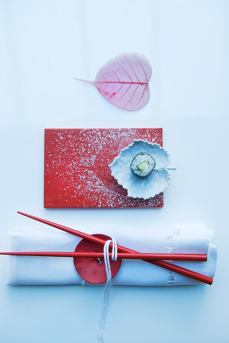 Asiatische Tischdeko: weiße Serviette mit grossem Knopf als Serviettenring, Essstäbchen und rote Platte und blattförmige Keramikschale mit Puderzucker verziert