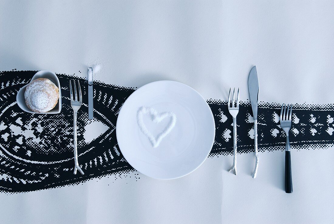 Weisser Teller mit Herzform aus Zuckerstreuseln und Besteck mit auffälligem Griff neben Gebäck in herzförmiger Schale auf Tapetenrolle mit schwarzer Zeichnung