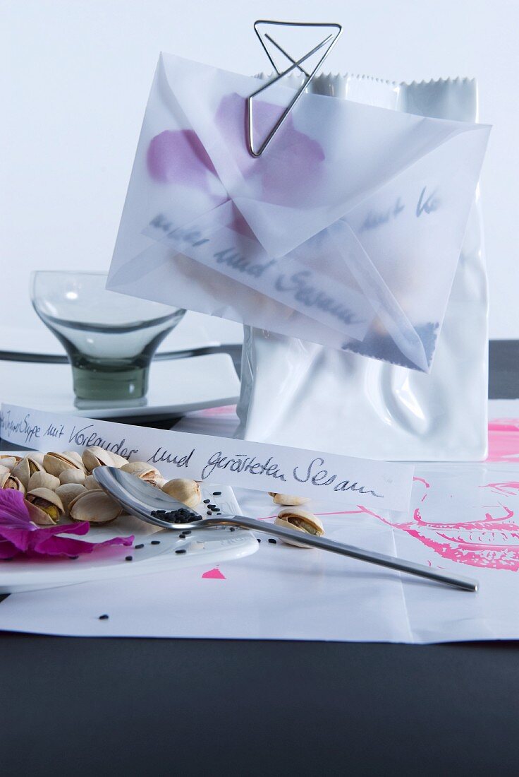 Translucent envelope filled with sesame seeds and petals clipped to vase shaped like paper bag with paper clip