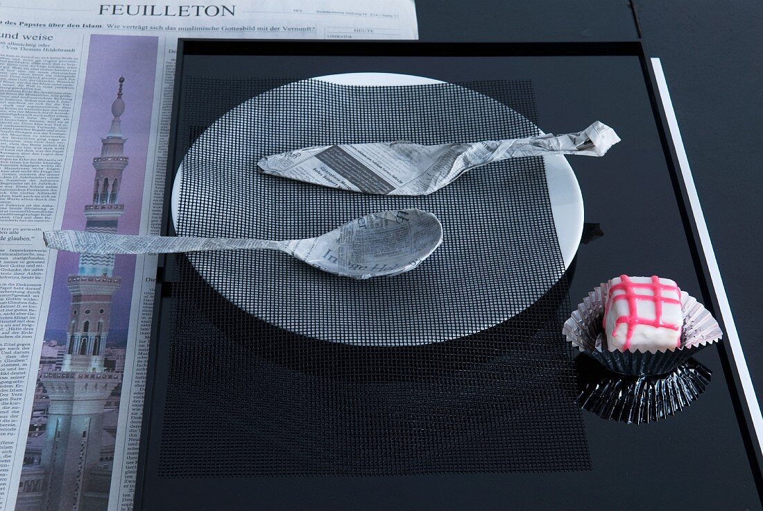 Mit Zeitungspapier umwickeltes Besteck auf Teller mit einem schwarzem Netz als puristische Tischdeko