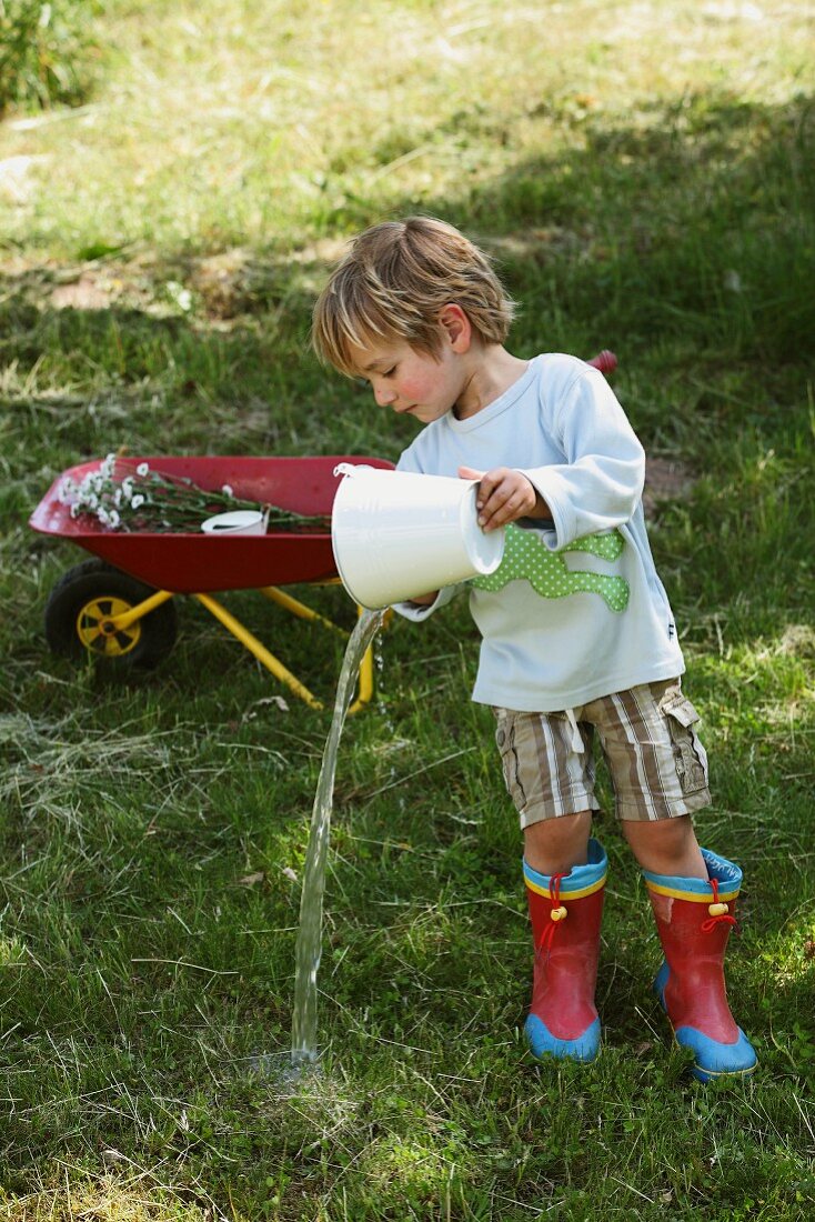 Kind in Gummistiefeln & mit selbstgenähtem Shirt spielt mit Wassereimer im Garten