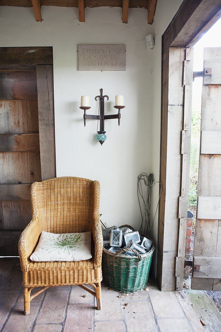 Wrought iron candle sconce above wicker chair next to basket on stone floor in rustic foyer with open front door