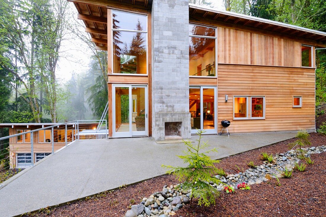 Gemauerter Kamin auf einer dreieckigen Terrasse vor modernem Holzhaus mitten im Wald