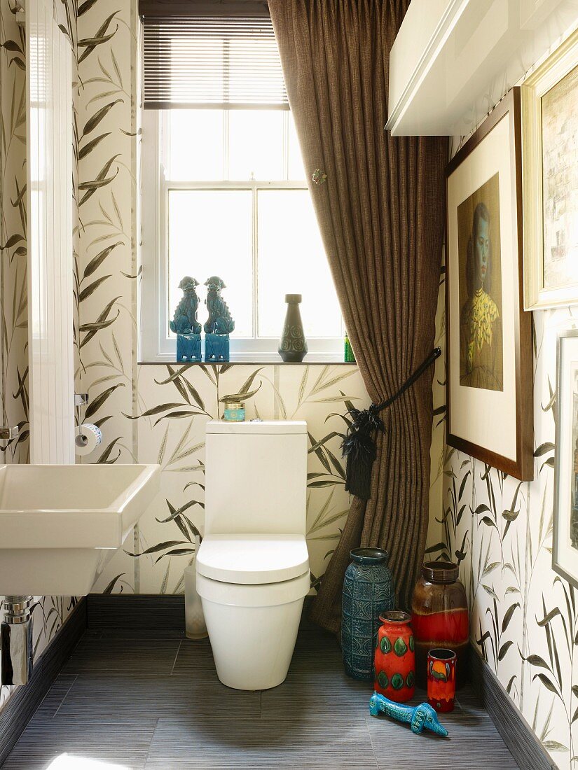 Large tiles with floral pattern in bathroom with draped curtains & collection of retro vases next to toilet