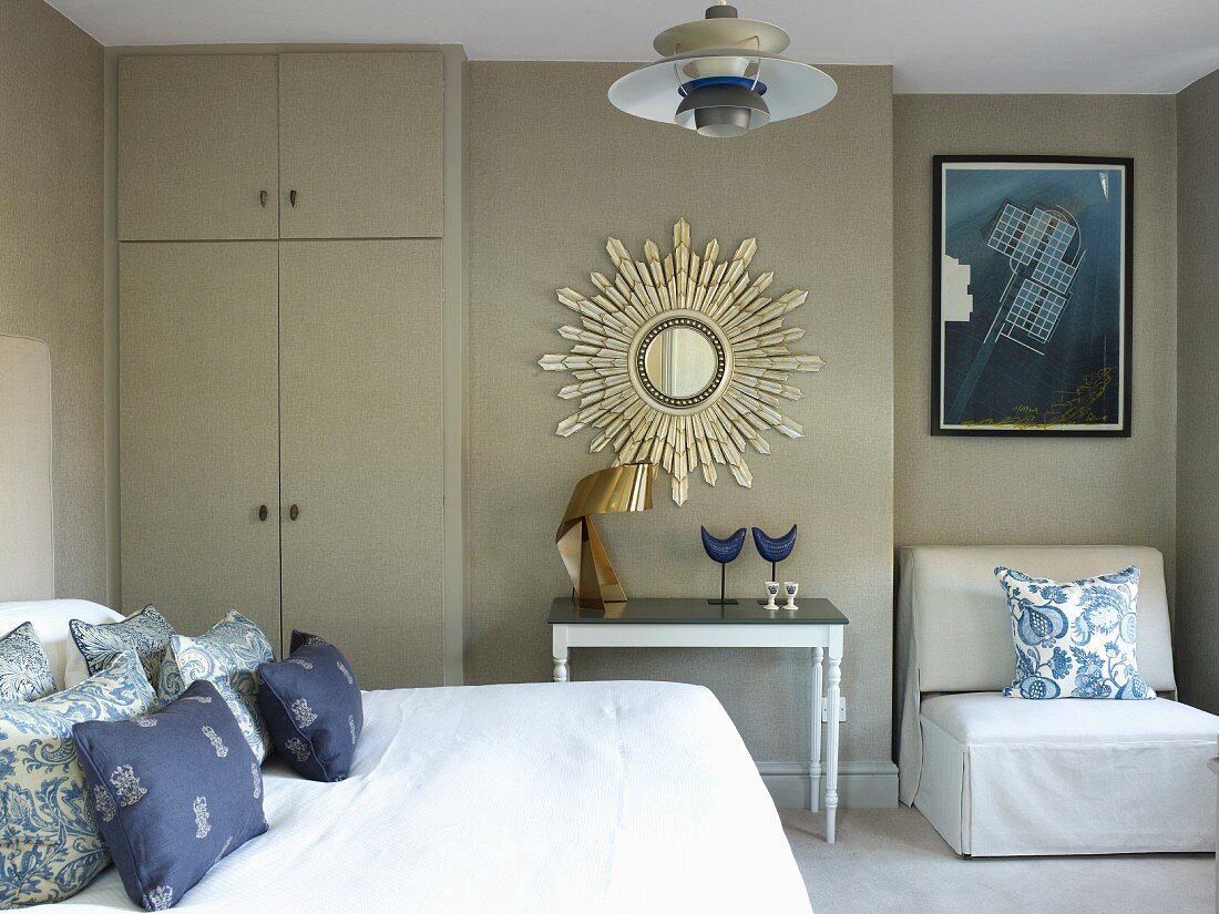 Schlafzimmer in Grau- & Blautönen mit Doppelbett, raumhohem Schrank, Spiegel mit strahlenförmigem Rahmen, Beistelltisch und Polstersessel in Nische