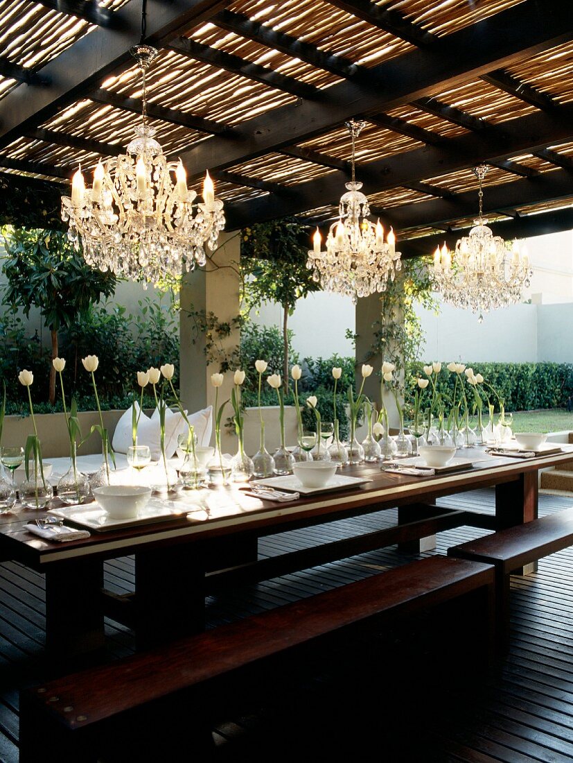 Outdoor Tisch mit integrierten Bänken auf überdachter Holzterrasse, festlich gedeckt mit einer Reihe weisser Tulpen unter Kronleuchtern