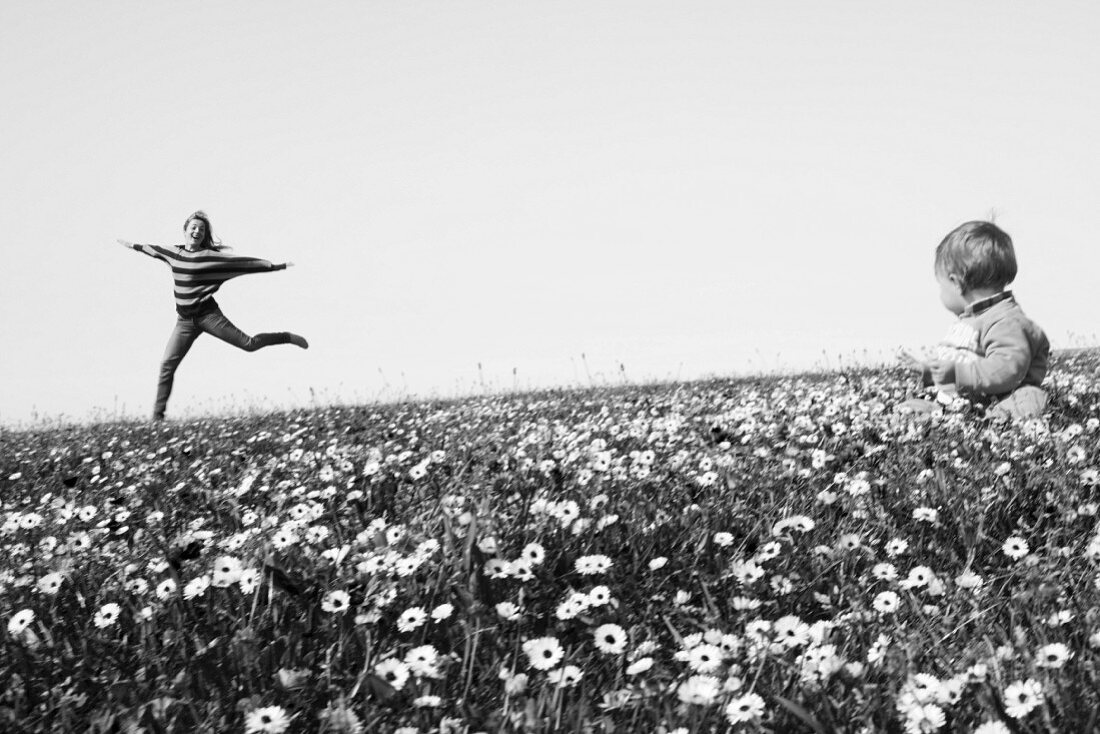 Frau springt fröhlich auf einer Blumenwiese, Baby sitzt im Vordergrund