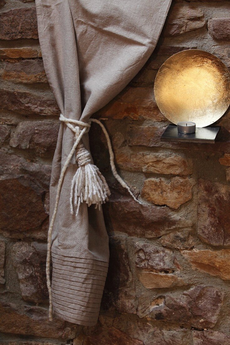 Vorhang mit Quaste aus Filzwollschnüren neben Windlicht und Metallschale an Natursteinwand