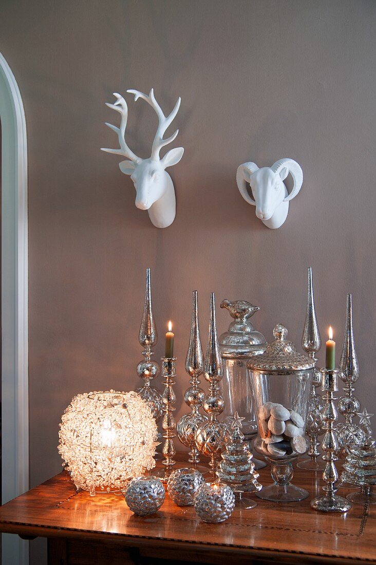 Kugelförmiges Windlicht neben Weihnachtsschmuck in Silber auf antikem Tisch, darüber Porzellantierköpfe an der Wand