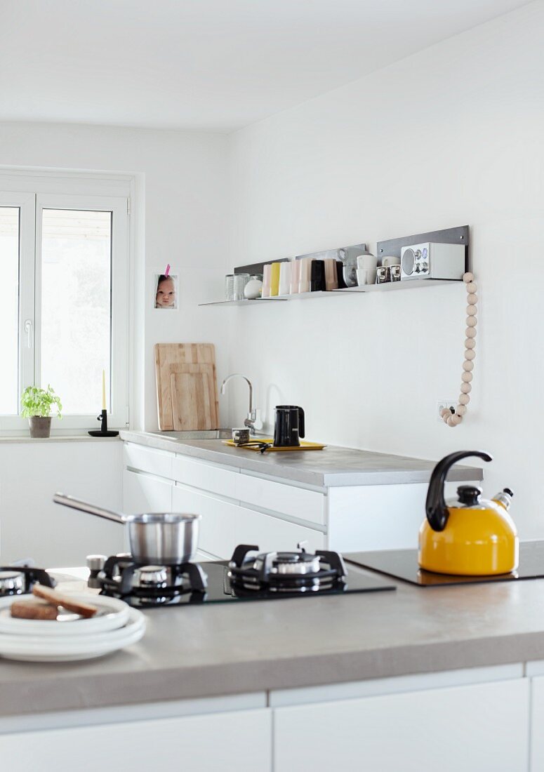 Gelber Wasserkessel auf Küchentheke mit Gaskochfeld in Betonarbeitsplatte, im Hintergrund Küchenzeile unter minimalistischer Wandkonsole aus Metall