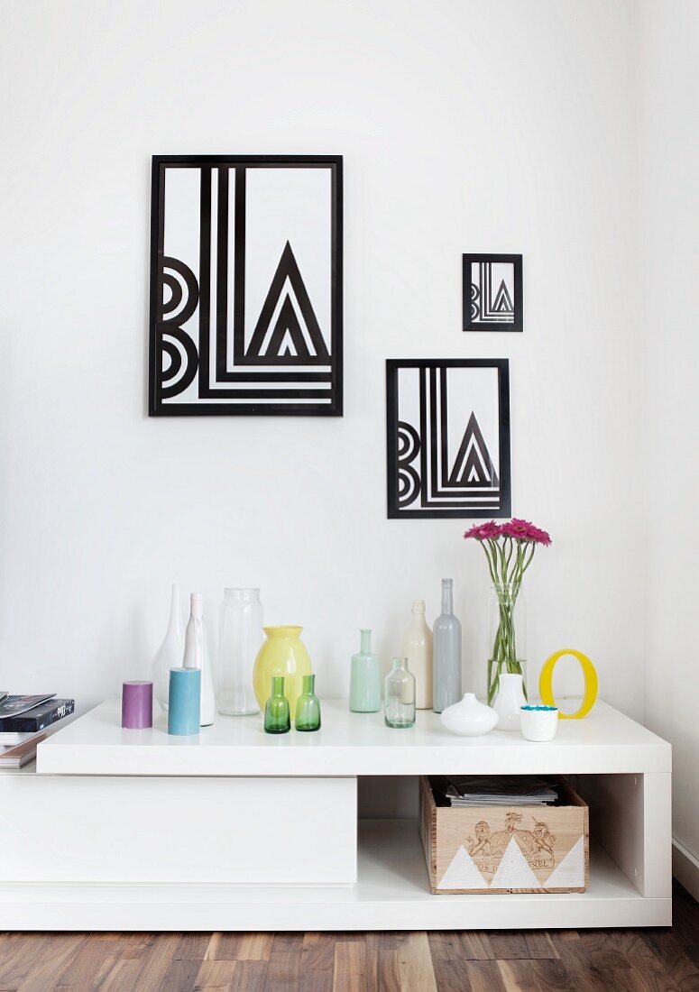 Collection of coloured bottles on white, low sideboard below framed, black and white, geometric graphics on wall