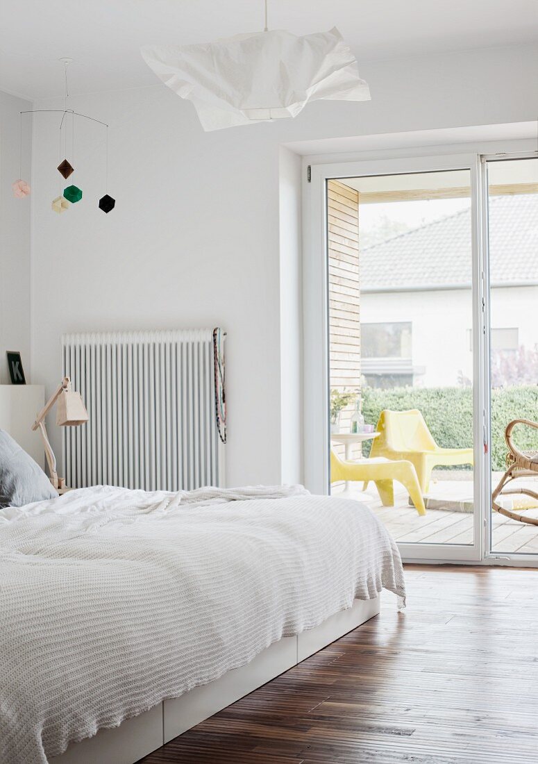 Doppelbett mit weisser Tagesdecke neben Terrassentür mit Ausblick; weiße Papierleuchte und Mobile an Decke in minimalistischem Schlafzimmer