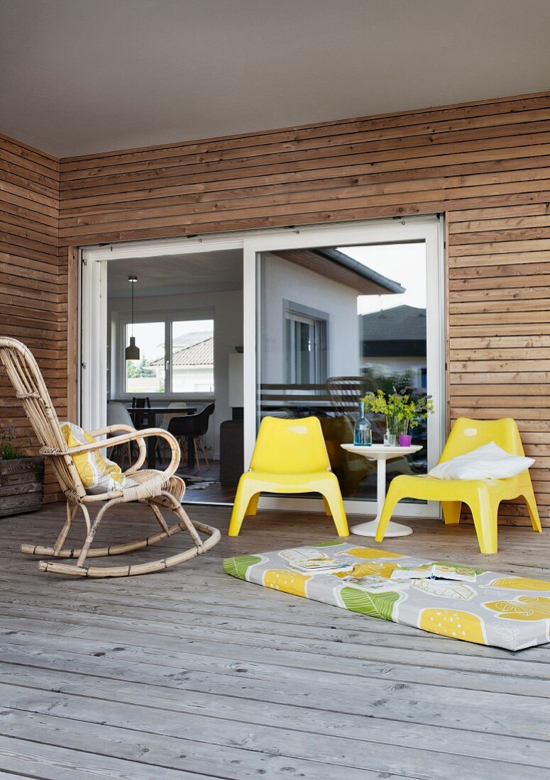 Wicker rocking chair, yellow plastic chairs and long cushion on floor of roofed wooden terrace