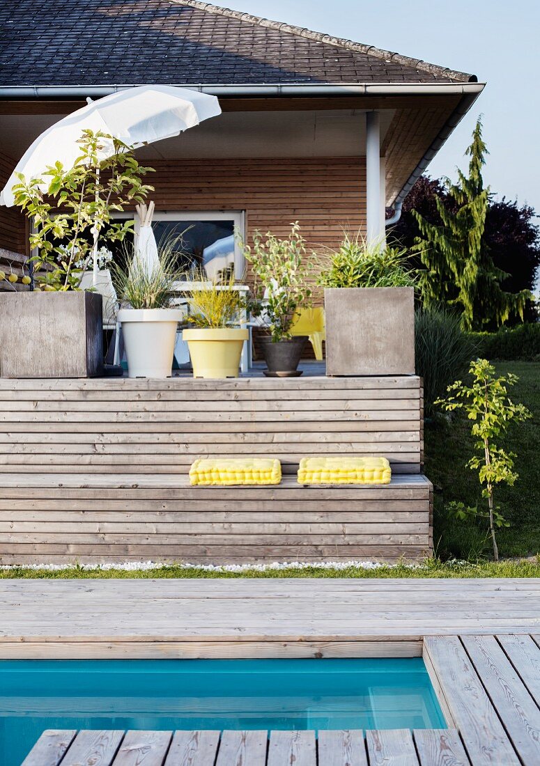 Modernes holzverschaltes Wohnhaus mit Pflanztöpfen und Sonnenschirm auf Veranda; vorgelagert Holzbank mit gelben Sitzpolstern und Pool mit umlaufender Holzterrasse