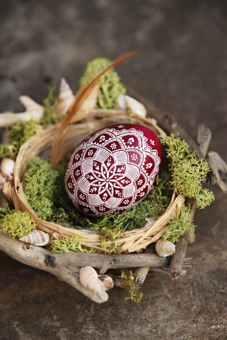 Kunstvoll mit weißem Spitzenmotiv bemaltes Ei in Nest aus Ästen und Moos mit Muscheldeko