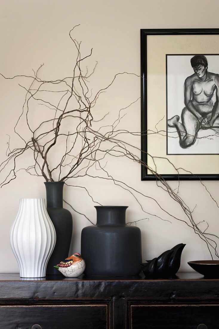 White and black vases of twigs on black Oriental sideboard below framed drawing of nude