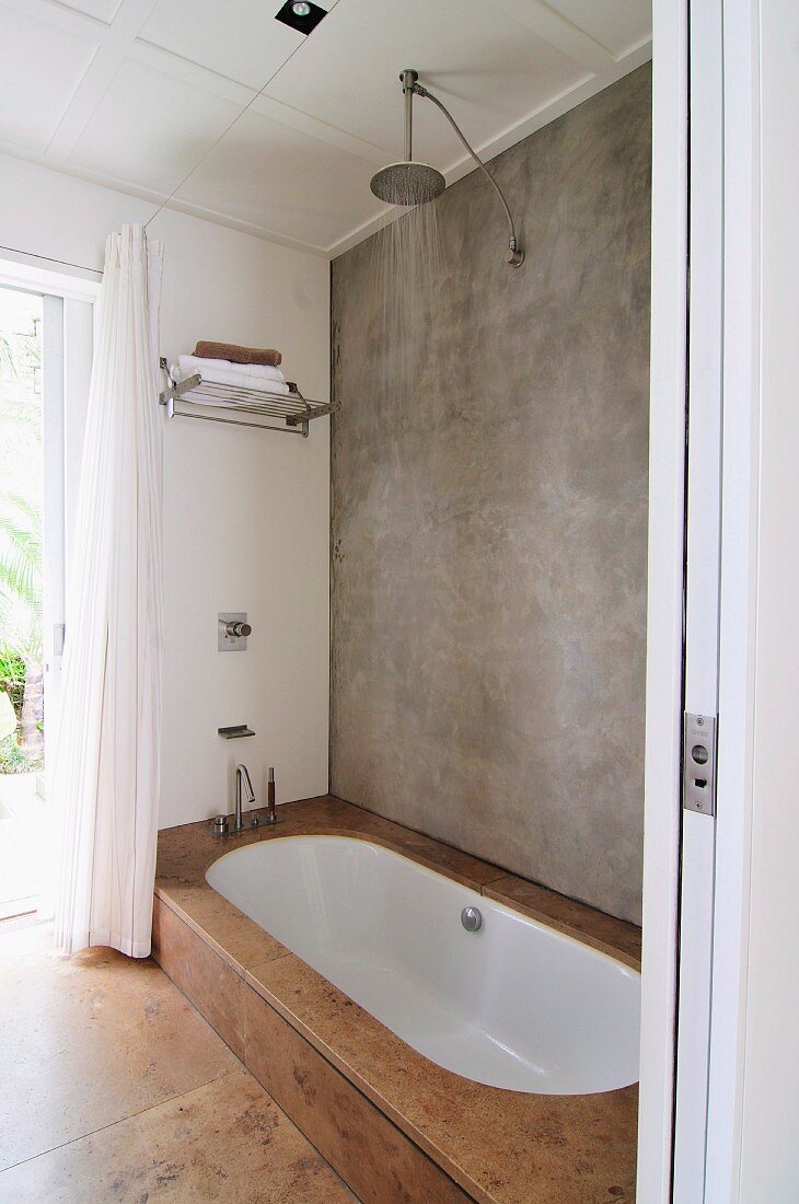 Concrete wall, rainfall shower and sunken white bathtub with stone-tiled surround in minimalist bathroom