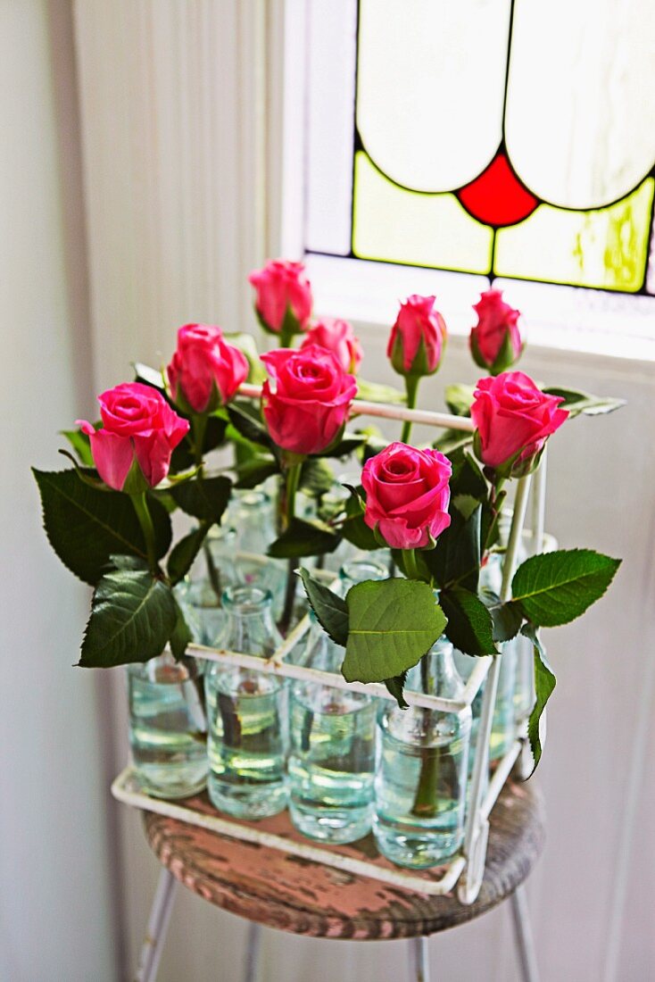 Pink roses in bottles in vintage metal bottle carrier on simple stool in front of Art Nouveau, stained-glass window