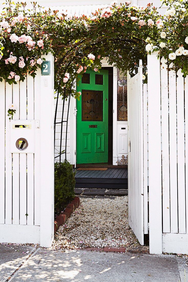 Weisslackierter Gartenzaun mit offener Tür und Torbogen mit berankten Kletterrosen vor grüner Haustür