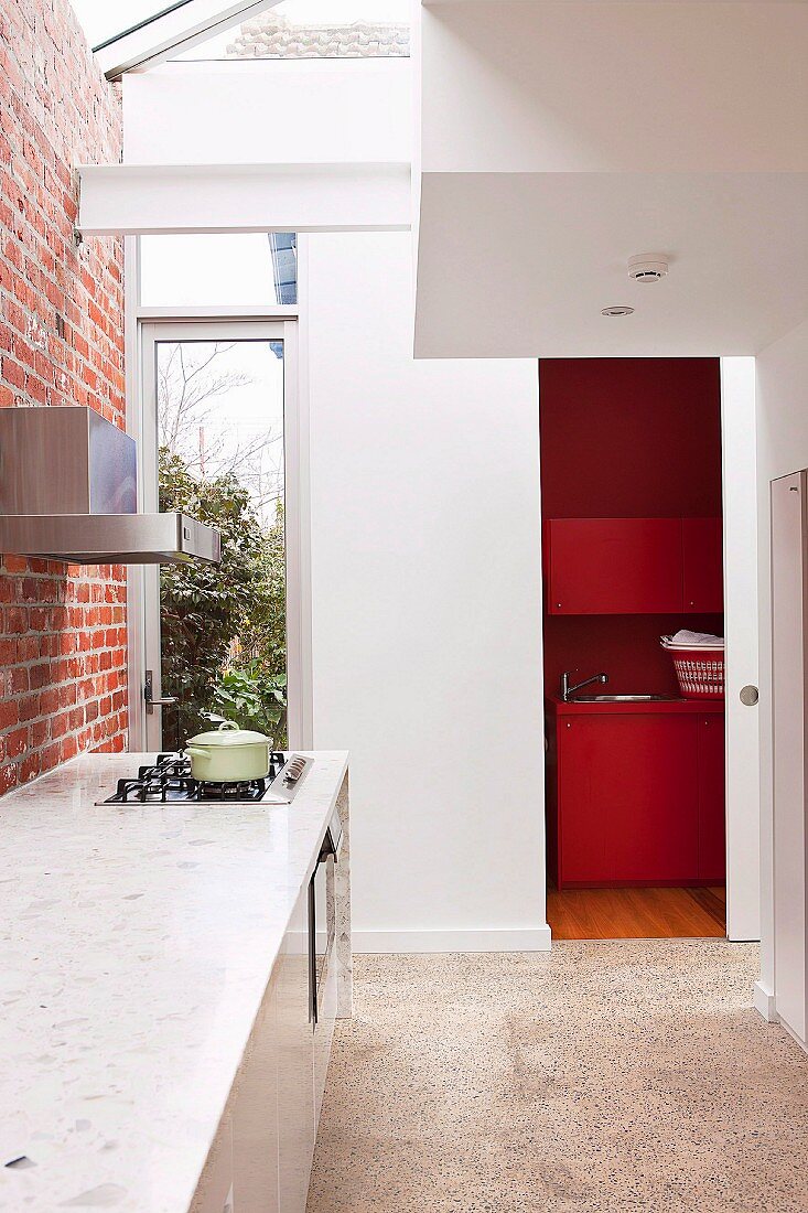Schmale, zeitgenössische Küche mit Oberlicht in Deckenbereich - an Ziegelwand Küchenzeile mit weisser Stein Arbeitsplatte, im Hintergrund offene Schiebetür und Blick ins rotgetönte Bad