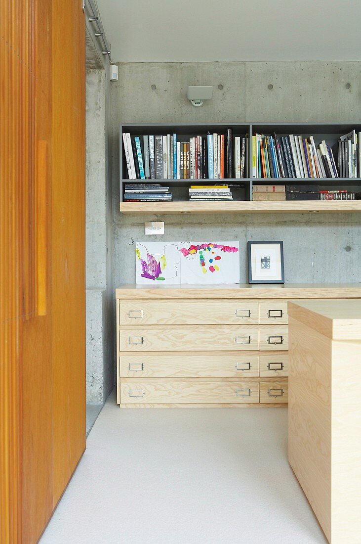 Ecke in modernem Wohnraum - halbhoher Schubladenschrank aus hellem Holz, oberhalb graues Wandregal an Sichtbetonwand, seitlich Schiebetür aus Holz