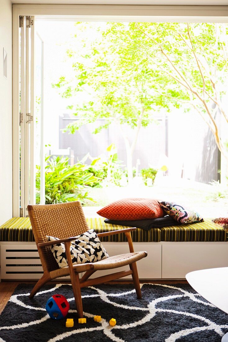 Comfortable retro wooden armchair on graphic black and white rug, open glass wall and view into green sunny garden