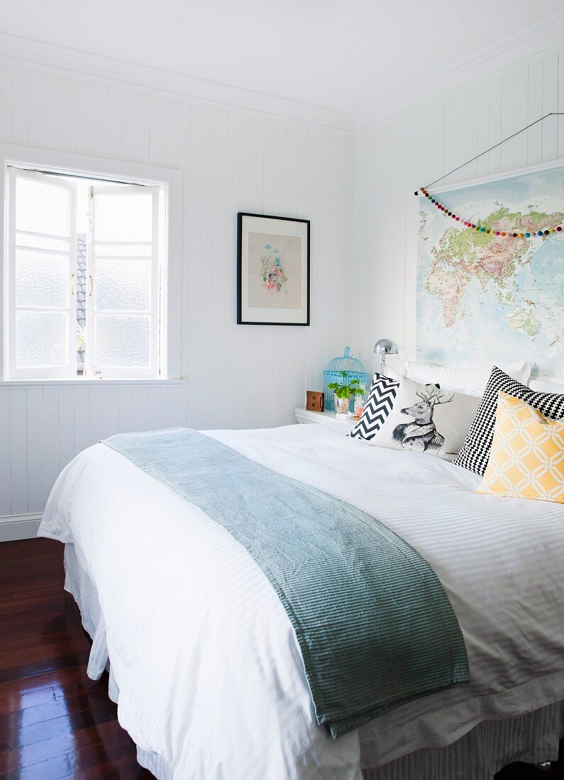 Bequemes Doppelbett mit verschiedenen Kissen, geöffnetes Sprossenfenster und aufgehängter Landkarte