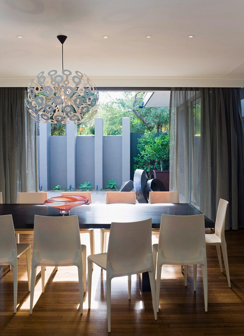 Kugelförmige Hängeleuchte Dandelion von Richard Hutten über Esstisch mit Designer-Stühlen aus hellem Kunststoff; Blick auf Terrasse mit blaugrauer Sichtschutzwand