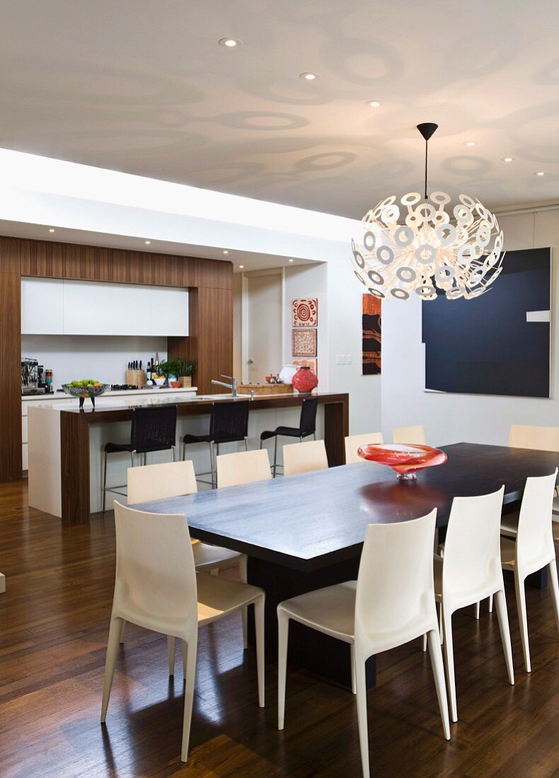 Esstisch mit Designer Hängeleuchte Dandelion und hellen Kunststoff Stühlen; offene Küche mit Frühstückstheke im Hintergrund