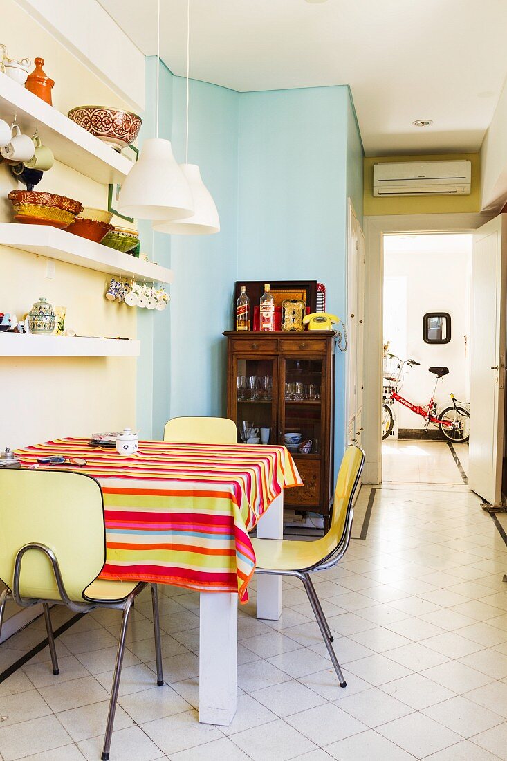Small, 50s-style dining area with table, brightly striped tablecloth and vintage chairs; bicycle in hallway in background