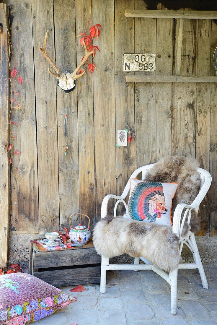 Rustikale Holzhüttenwand mit Geweih dekoriert und verwitterte Holzkiste als Teetischchen neben gemütlichem Rattansessel mit kuscheligem Schaffell