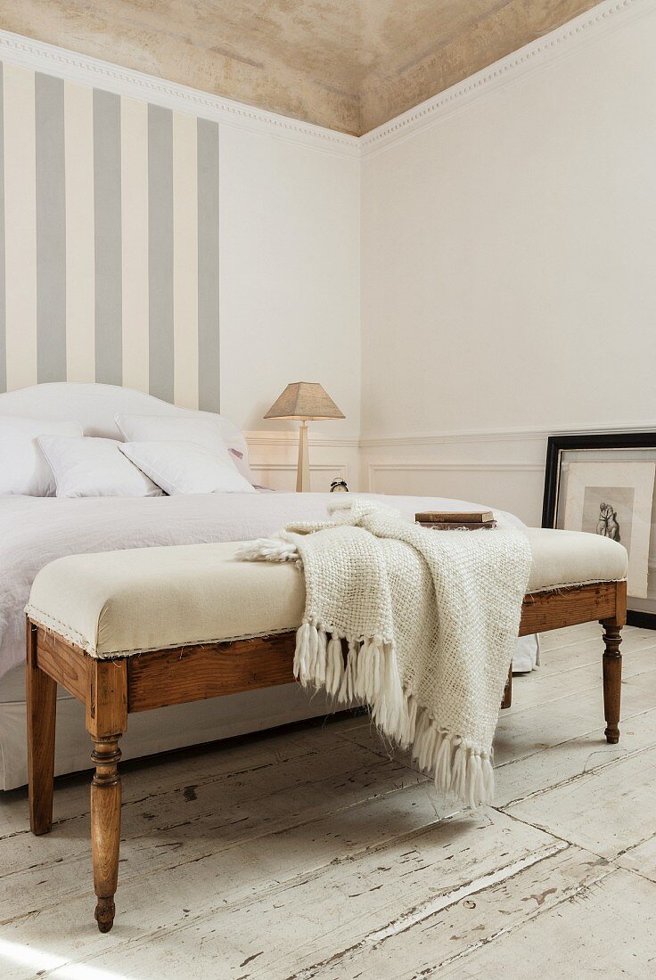 Strickplaid auf gepolsterter Bank an Bettende eines französischen Bettes und grau-weisses Streifenmuster an Wand